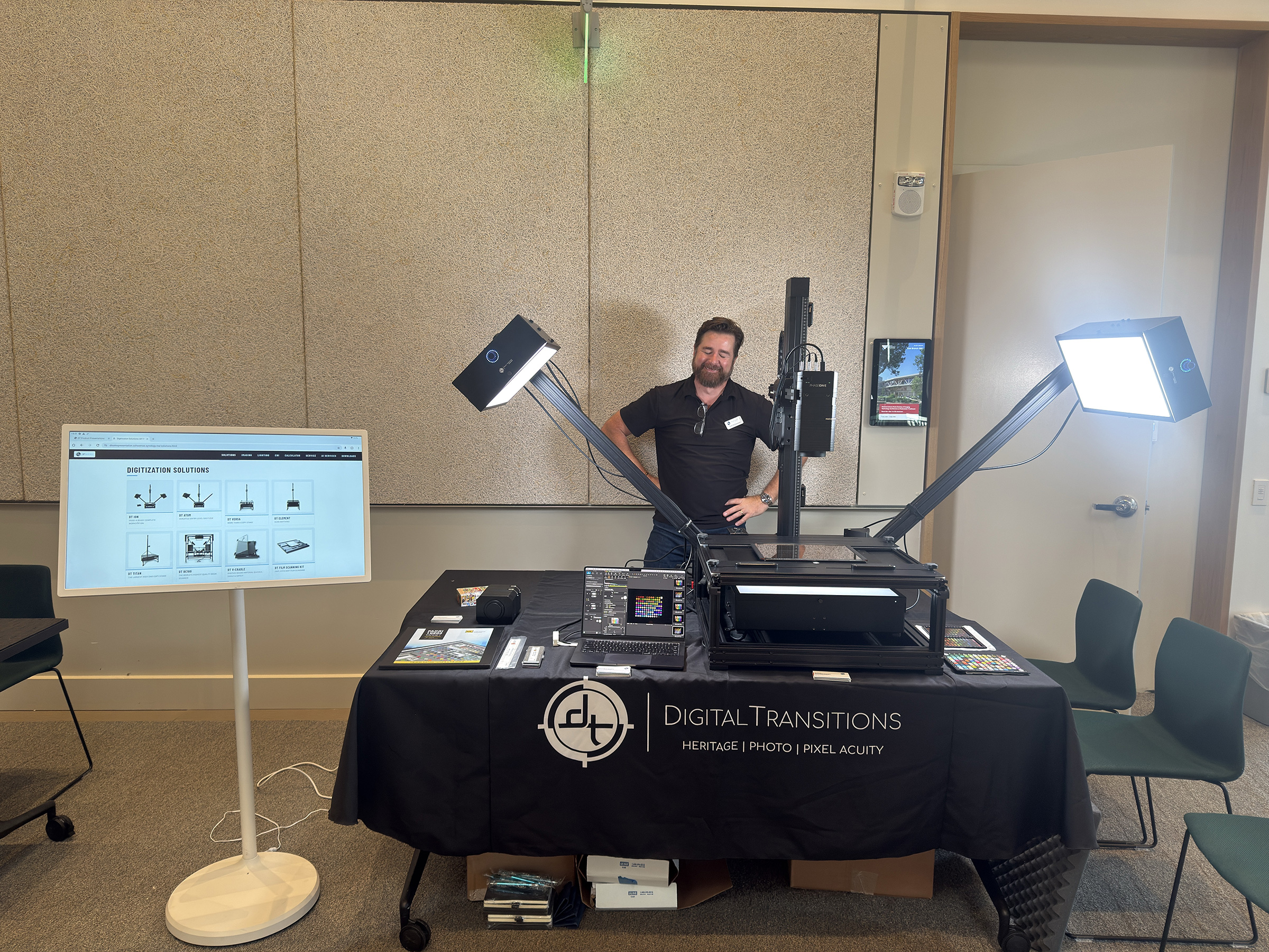 The Digital Transitions exhibit table at Archiving 2026, with a Phase One capture system on a DT Atom stand, Stellar MKII lighting, and a monitor cycling through DT's digitization solutions